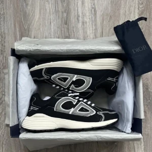 Svarta Dior sneakers med vita detaljer - Snygga svarta sneakers från Dior med stora CD-loggor i vitt och grått på sidorna. Skorna har en chunky vit sula och vita skosnören. Tillverkade i en mix av textil och syntetmaterial för en modern look.