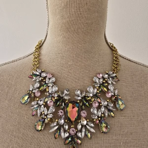 Statement halsband med färgglada stenar - Ett riktigt statement halsband med guldtonad kedja och stora glittriga stenar i regnbågsfärger, rosa och klara kristaller. Blommiga former och droppformade detaljer ger en lyxig och unik look. Perfekt för dig som vill sticka ut och glänsa.