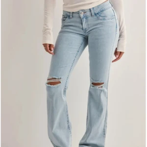 Ljusblå bootcut jeans med slitningar - Säljer ett par ljusblå bootcut jeans med slitna hål över knäna och snygga broderade detaljer i rött på bakfickorna. Jeansen har klassisk femficksdesign, normal passform och är tillverkade i mjukt denimtyg. Perfekta för en avslappnad och trendig look. Lite slitna nere på jeansen. Jeansen är från Nelly. 
