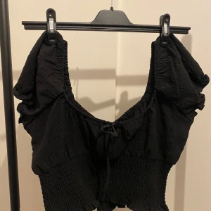 Svart croppad blus från H&M - Svart croppad blus från H&M med puffiga korta ärmar och smockad midja. Toppen har en söt knytning framtill och volangkant nertill. Tillverkad i mjuk polyester med lite stretch för extra komfort. Perfekt för dig som gillar trendiga och feminina detaljer.