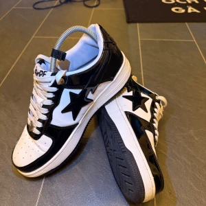 Svarta och vita Bape Sta sneakers - Säljer ett par Bape Sta sneakers i svart och vitt med glansiga svarta paneler och den ikoniska stjärnan på sidan. Skorna har vita snören, perforerad tå och låg profil. Klassisk streetwear-vibe och riktigt snygga detaljer som metallplakett på snörningen.