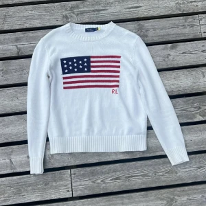 Vit stickad tröja Polo Ralph Lauren - Vit stickad tröja från Polo Ralph Lauren med amerikansk flagga i blått och rött på bröstet samt RL-broderi. Tröjan har rund hals, ribbade muddar och långa ärmar. Perfekt för dig som gillar klassisk och stilren design med en cool detalj framtill.