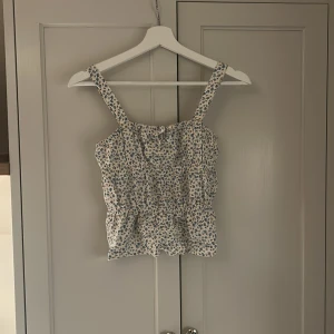 Blommig linnetopp med volang - Supersöt linnetopp i vitt med små blå blommor och volangkant. Toppen har breda axelband och smockad midja som ger en avslappnad och somrig vibe. Perfekt till jeans eller kjol för en fräsch look.