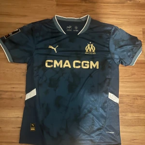 Marseille matchtröja Puma 24/25 - Snygg Olympique de Marseille fotbollströja från Puma i mörkblått med guldiga detaljer och klubbmärke. Kortärmad modell med ljusblå och vita inslag vid krage och ärmslut. Använd 1/2 gånger, används inte då den inte passar mig. Pris kan diskuteras vid snabb affär.
