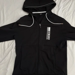 Svart vindjacka från BOSS med huva - Svart vindjacka från BOSS med vit logga på bröstet och snygga vita detaljer på huvan. Jackan har dragkedja framtill, huva med snörning och två fickor. Perfekt för dig som gillar stilren och sportig look.