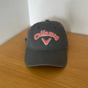 Callaway golf keps  - Snygg mörkgrå golf keps från Callaway med broderad logga i rött och vitt framtill. Justerbar rem baktill med metallspänne och röd Callaway-detalj. Tillverkad i 100% bomull, perfekt för golf eller chill vardagsstil.
