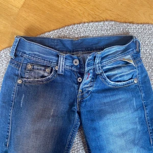 Blå Replay jeans med broderad patch - Snygga blå jeans från Replay med slitningar och coola detaljer på bakfickorna. De har en broderad gul patch med blommor bak och klassisk femficksdesign. De är uppsprättade längst ner. Skriv om du vill ha fler bilder. Midjan rakt över: 34 cm. Innerben: 75cm.