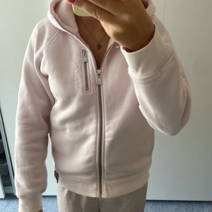 Ljusrosa hoodie från Sail Racing - Ljusrosa hoodie från Sail Racing med dragkedja framtill och extra zip-detalj på bröstet. Stor logga på ryggen och broderad text på framsidan. Mjuk bomullskänsla, huva och långa ärmar. Perfekt för chill dagar och snygg att matcha med jeans eller mjukis.