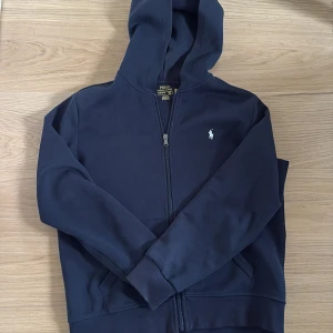 mörk blå hoodie från Polo Ralph Lauren - Svart hoodie från Polo Ralph Lauren med dragkedja och klassisk huva. Tröjan har det ikoniska broderade logot på bröstet och ribbade muddar. Perfekt för en chill och stilren look. säljs för att den är för liten.
