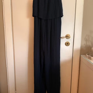 Mörkblå plisserad byxdress från ICHI - Supersnygg mörkblå byxdress från ICHI i plisserat tyg. Vida ben och flowig passform som ger en chill och trendig vibe. Perfekt för dig som gillar stilrena och bekväma plagg med det lilla extra. Resår i midjan och mjukt, lätt material. ( Har aldrig använts)