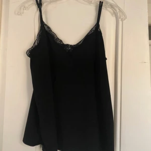 Svart linnetopp med spetsdetalj - Snygg svart linnetopp med tunna axelband och v-ringning dekorerad med spets. Perfekt för att styla lager på lager eller bära som den är. 