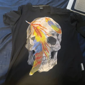 Svart t-shirt med döskalle från Philipp Plein - Svart t-shirt från Philipp Plein i bomull med stor döskalleprint framtill, dekorerad med färgglada fjädrar i rött, gult och blått. Rund hals och korta ärmar. Snygg statement-piece för dig som gillar edgy stil och coola detaljer.