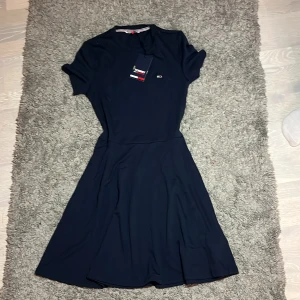 Marinblå kortklänning Tommy Hilfiger - Snygg marinblå kortklänning från Tommy Hilfiger med rund hals och korta ärmar. Aldrig använd Klänningen har en enkel, stilren design med ett litet broderat logomärke på bröstet. Materialet är mjukt och följsamt, perfekt för dig som gillar klassisk och clean stil.