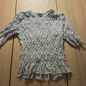 Prickig blus med volang från Gina Tricot💖 - Supersöt vit blus från Gina Tricot i storlek XS, med svarta prickar och smockad struktur över hela plagget. Blusen har trekvartsärm och volangkant nedtill som ger en extra feminin touch. Perfekt för dig som gillar trendiga detaljer och mönster.