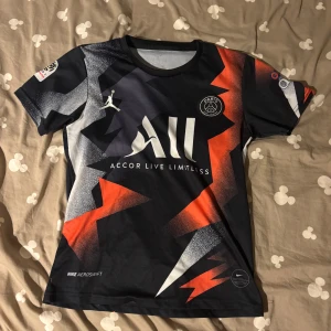 PSG Neymar Jr 10 matchtröja Nike - Paris Saint-Germain fotbollströja med Neymar Jr och nummer 10 på ryggen. Svart bas med skarpa röda, vita och gråa blixtmönster. Kortärmad, tillverkad i lätt och ventilerande material med Jordan-logga och Accor Live Limitless tryck på bröstet. Snygg och modern design för dig som älskar fotboll.