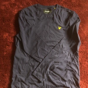 Mörkblå långärmad tröja Lyle & Scott - Snygg mörkblå långärmad tröja från Lyle & Scott med klassisk rund hals och gul broderad logga på bröstet. Tröjan är tillverkad i mjuk bomull och har en clean, enkel design som passar perfekt till jeans eller chinos.