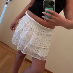 Vit blommig volangkjol med resår - Supersöt kort kjol i vitt med blått blommönster och volanger. Kjolen har bred resår i midjan för skön passform och är gjord i ett lätt, luftigt material som känns perfekt för varma dagar. Den har flera lager och ger en romantisk vibe.