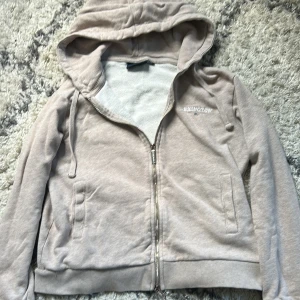 Beige hoodie från Lexington med dragkedja - Säljer en beige hoodie från Lexington med huva och dragkedja framtill. Tröjan har långa ärmar, ribbade muddar och två fickor på sidorna. Mjuk bomullskvalitet och diskret Lexington-logga på bröstet. Perfekt för chill dagar eller när du vill vara extra comfy.