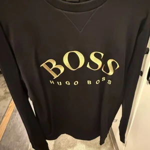 Svart långärmad tröja Hugo Boss - Svart långärmad tröja från Hugo Boss med stort gult BOSS-tryck på bröstet. Klassisk rund halsringning och stilren design som passar perfekt till jeans eller dressade byxor. Skön och enkel att matcha för en clean look.