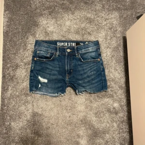 Blå jeansshorts med rå kant - Snygga blå jeansshorts från Super Stretch med klassisk femficksdesign och rå, fransig kant nedtill. Shortsen har slitna detaljer framtill och en brun patch bak i midjan. Perfekta för sommarens chill dagar och har en normal passform.