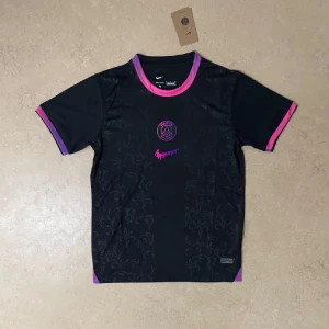 PSG x Nike svart fotbollströja - Svart PSG fotbollströja från Nike med diskret blommönster i tyget. Rosa och lila detaljer vid krage och ärmslut, samt rosa Nike-logga och PSG-emblem på bröstet. Tillverkad i lätt och ventilerande material, perfekt för träning eller match.