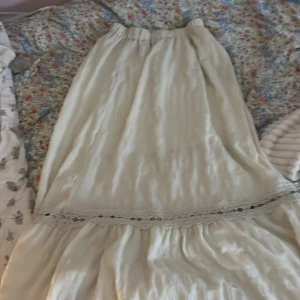 Vit långkjol med spetsdetalj - Säljer en superfin vit långkjol med elastisk midja och volang. Kjolen har en bred spetsdetalj i midjan som ger en romantisk vibe. Materialet är lätt och har en skrynklig struktur, med ett mjukt foder på insidan för extra komfort.