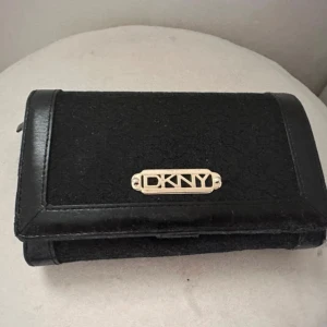Svart DKNY plånbok - Snygg svart plånbok från DKNY med guldfärgad logga framtill. Plånboken har flera fack för kort, myntfack med dragkedja och transparent fönster för ID eller bild. Utsidan är i en mix av skinn och textil