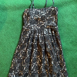 Svart spetsklänning från Vila - Supersnygg svart spetsklänning från Vila med axelband och blommigt spetsmönster över en ljus, silkeslen underklänning. Klänningen har en utställd kjol och markerad midja som ger en feminin siluett. Perfekt för dig som vill sticka ut med stil.