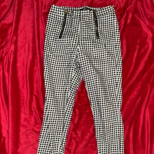 Svartvita rutiga byxor med dragkedjor - Snygga svartvita byxor med rutigt mönster och två coola dragkedjor framtill. Byxorna har smal passform och är perfekta för dig som vill sticka ut lite extra. De är midjehöga och gjorda i ett mjukt bomullsmaterial som känns skönt hela dagen.