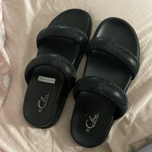 Svarta sandaler med glittriga band - Snygga svarta sandaler från din sko/Clou med två breda band täckta av glittriga detaljer. Platt sula och öppen tå gör dem perfekta för varma dagar. Stilren och trendig design som passar till många outfits.