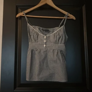 Rutig linnetopp från Brandy Melville - Supersöt svartvit rutig linnetopp från Brandy Melville med smala axelband, knappar framtill och dekorativ spetskant vid urringningen. Toppen har knytband i ryggen och en lätt utställd passform som ger en avslappnad vibe. Perfekt för varma dagar och enkel att styla.