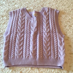 Ljuslila stickad väst från Kappahl - Superfin ljuslila stickad väst från Kappahl med grova flätstickade detaljer och v-ringad hals. Perfekt att slänga över en skjorta eller t-shirt för en trendig lager-på-lager-look. Mjuk och mysig känsla, ribbade kanter för extra stil.