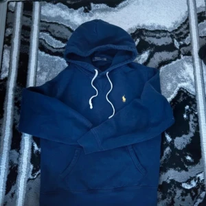 Polo Ralph Lauren hoodie  - Säljer en marinblå hoodie från Polo Ralph Lauren med klassisk gul broderad logga på bröstet. Tröjan har huva med vita snören och känguruficka framtill. Tillverkad i mjuk bomullsmix för extra komfort. Perfekt för en chill och stilren look.
