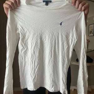 Vit långärmad tröja från Ralph Lauren - Klassisk vit långärmad tröja från Ralph Lauren med den ikoniska blå broderade loggan på bröstet. Tröjan är i mjuk bomull och har en enkel, stilren design som passar till det mesta. Perfekt basplagg för garderoben.