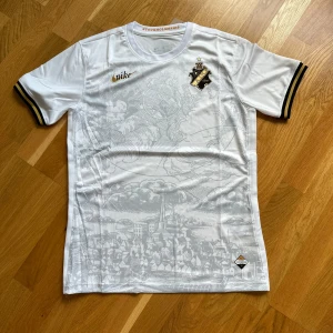 Vit AIK fotbollströja Nike Stockholm - Snygg vit AIK fotbollströja från Nike med guldsvart klubbmärke på bröstet och detaljerade tryck av Stockholms stad i grått framtill. Kortärmad med svarta och gula ränder på ärmsluten. Lätt och ventilerande material, perfekt för match eller träning.