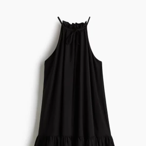 Svart omlott kortklänning med volang - Supersnygg svart kortklänning med omlott och volangdetaljer längst ner. Klänningen har en hög hals med snörning och är ärmlös. aldrig använd!