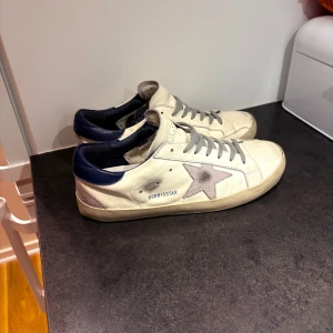 Golden Goose Superstar sneakers vit/grå - Golden Goose Superstar sneakers i vitt skinn med grå mockastjärna på sidan, grå snörning och mörkblå häl. Klassisk låg modell med rund tå och platt sula. Perfekta för en avslappnad och trendig streetstyle. Pris kan diskuteras!