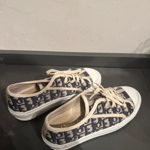  Dior sneakers med monogram - Snygga sneakers från Dior i blått och vitt med ikoniskt monogrammönster. Skorna har vita snören, rund tå och platt sula. Ovandelen är i textil med tydliga Dior-detaljer och logga på plösen. Perfekta för dig som vill sticka ut med en lyxig touch.