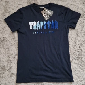 Trapstar mörkblå t-shirt med broderi - Snygg mörkblå t-shirt från Trapstar med broderad logga i olika blå nyanser och texten 'IT'S A SECRET' på bröstet. Klassisk rund hals och korta ärmar. Perfekt för dig som vill sticka ut med streetwear-vibe. Den passar Xs och S
