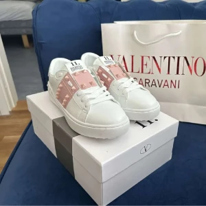 Valentino rosa sneakers - Du får med påse å skorna! Snygga Valentino Garavani Rockstud Open sneakers i vitt skinn med breda ljusrosa band och ikoniska nitar. Klassisk rund tå, platt sula och vita snören. Perfekta för dig som vill sticka ut med exklusiva detaljer och modern design. Storlek:39. Använd en gång. Säljer för de är för små för mig