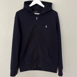 Mörkblå hoodie från Ralph Lauren - Klassisk svart hoodie från Ralph Lauren med dragkedja framtill och vit broderad logga på bröstet. Tröjan har huva, känguruficka och långa ärmar. Perfekt för en chill och stilren look.