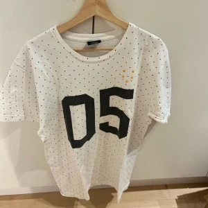 Vit prickig t-shirt från Undefeated - Vit t-shirt från Undefeated med svarta prickar, stor svart 05-tryck på framsidan och texten 'RUTHLESS' på ryggen. Tre små gula stjärnor på bröstet. Skön passform och klassisk rund hals. Perfekt för dig som gillar streetwear och sportig vibe.