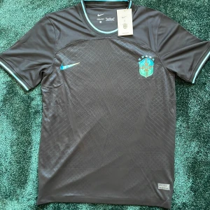 Mörkgrå Brasilien Nike fotbollströja - Snygg mörkgrå Brasilien fotbollströja från Nike med korta ärmar och diskret rutmönster. Tröjan har ett blått Brasilien-emblem och blå Nike-logga på bröstet. Tillverkad i lätt, ventilerande polyester med ribbade detaljer vid krage och ärmslut.