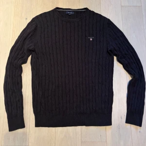 Svart stickad tröja från GANT - Säljer en svart stickad tröja från GANT i storlek L. Tröjan har ett klassiskt kabelstickat mönster, rund hals och ribbade muddar vid ärmslut och nederkant. Perfekt för dig som gillar stilrena och tidlösa plagg med diskret logga på bröstet.