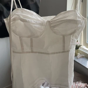 Vit korsett-topp med tunna axelband - Snygg vit korsett-topp med tunna axelband och markerade kupor. Toppen har en lätt transparent känsla och figurnära passform, perfekt för att skapa en trendig look. Materialet känns luftigt och passar bra till varma dagar.