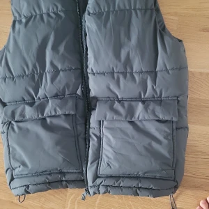 Grå dunväst från lager 157 stl xs - Snygg grå dunväst från New Garments i storlek XS. Västen har hög krage, två stora fickor framtill med lock och dragkedja hela vägen. Perfekt att slänga över en hoodie för extra värme. Enkel och stilren design som funkar till många outfits.
