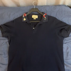 Svart Gucci t-shirt med broderad krage - Snygg svart t-shirt från Gucci med korta ärmar och unik krage med broderade detaljer som orm och bi. Klassisk pikémodell med knappar framtill. Perfekt för dig som vill sticka ut med exklusiva detaljer och streetstyle-vibe.
