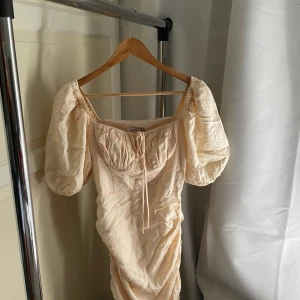 Champagnefärgad kortklänning från Adoore - Supersöt kortklänning från Adoore i champagnefärg med puffärmar och smockad byst. Klänningen har knytning framtill och en lätt rynkad effekt längs kjolen. Perfekt för sommarens alla tillfällen!