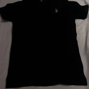 Svart pikétröja från U.S. Polo Assn. - Klassisk svart pikétröja från U.S. Polo Assn. med broderad logga på bröstet. Tröjan har korta ärmar, krage och knappar framtill. Perfekt för en clean och sportig look. Materialet är mjukt och andas, troligtvis bomull.