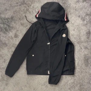 Svart vindjacka från Moncler med huva - Säljer en svart vindjacka från Moncler med huva och dragkedja. Jackan har Moncler-logga på bröstet och rödvit rand på huvan. Perfekt för blåsiga dagar och enkel att matcha med det mesta.